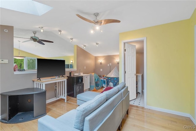 living area featuring a ceiling fan, light wood-style flooring, vaulted ceiling, a skylight, and baseboards