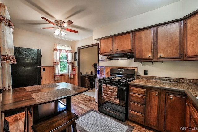 kitchen with black appliances, wood finished floors, a ceiling fan, wainscoting, and dark countertops