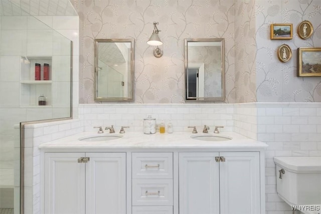 bathroom with wallpapered walls, double vanity, wainscoting, tile walls, and toilet