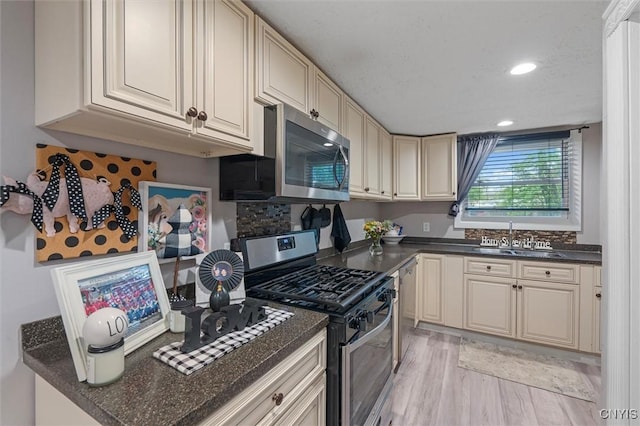 kitchen featuring stainless steel appliances, a sink, cream cabinets, dark countertops, and light wood-style flooring