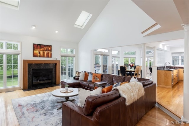 living room with decorative columns, high vaulted ceiling, a fireplace with flush hearth, light wood-style floors, and a skylight