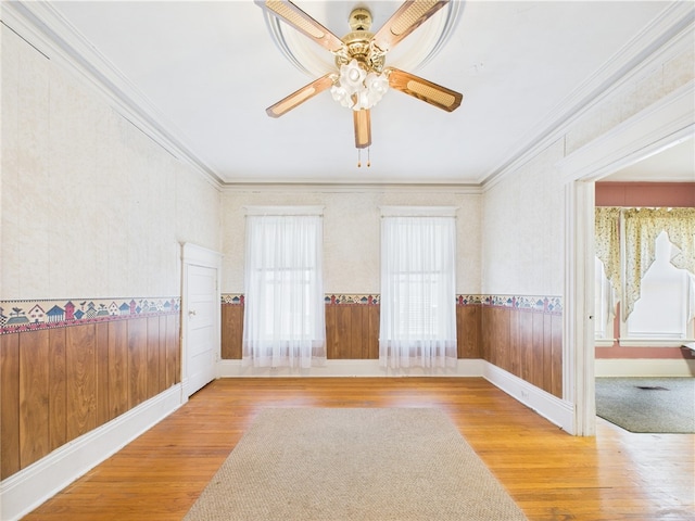 spare room with ornamental molding, wood finished floors, wainscoting, a ceiling fan, and wallpapered walls