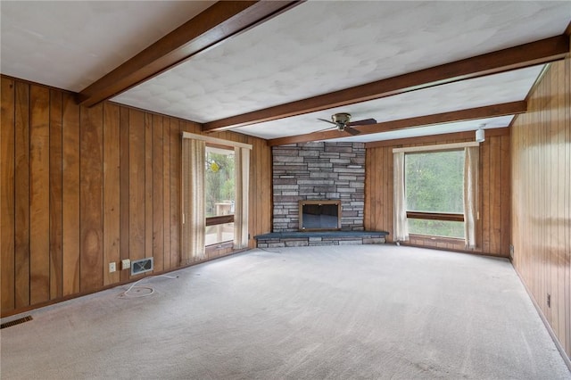 unfurnished living room featuring wooden walls, beamed ceiling, plenty of natural light, a fireplace, and carpet flooring