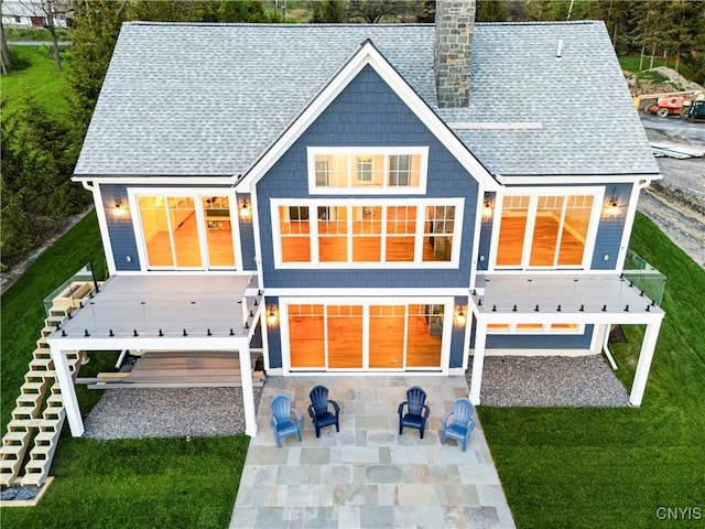 back of house featuring a chimney, a patio, a shingled roof, a wooden deck, and a lawn