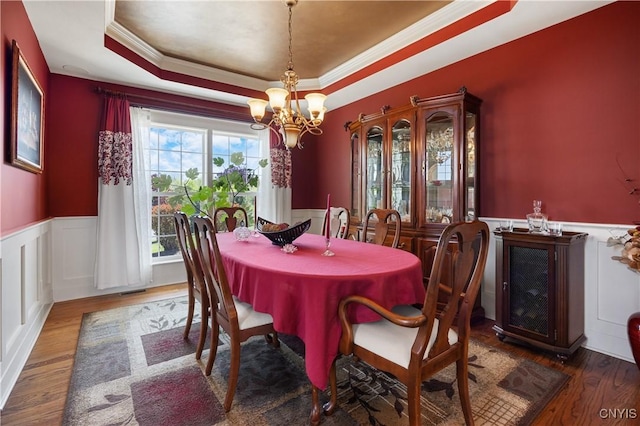 dining area featuring a raised ceiling, a chandelier, a wainscoted wall, ornamental molding, and wood finished floors