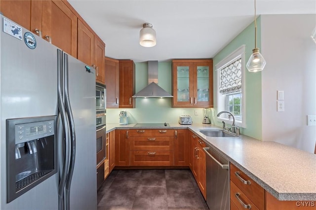 kitchen featuring appliances with stainless steel finishes, a sink, wall chimney exhaust hood, brown cabinets, and glass insert cabinets