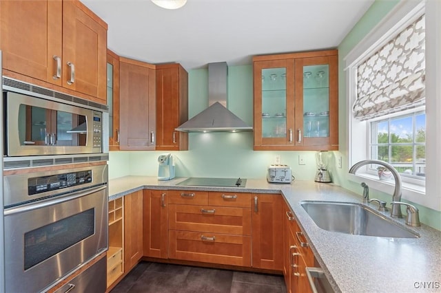 kitchen with stainless steel appliances, wall chimney exhaust hood, a sink, glass insert cabinets, and brown cabinets