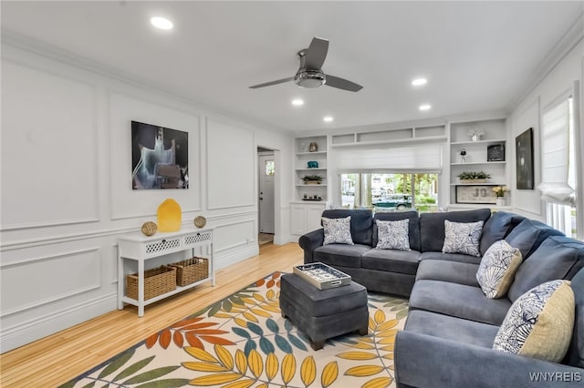 living room featuring light wood-type flooring, ornamental molding, a decorative wall, a ceiling fan, and recessed lighting
