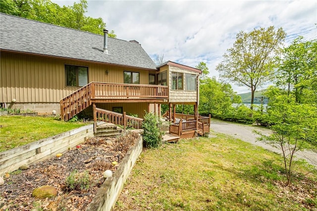 back of house featuring roof with shingles, a wooden deck, a sunroom, stairs, and a lawn