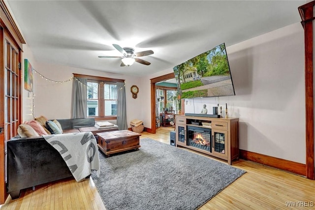 living area featuring baseboards, a ceiling fan, a glass covered fireplace, and light wood-style flooring