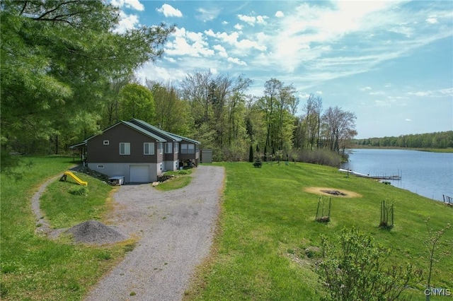 view of grassy yard featuring a water view, an attached garage, driveway, an outdoor fire pit, and a forest view