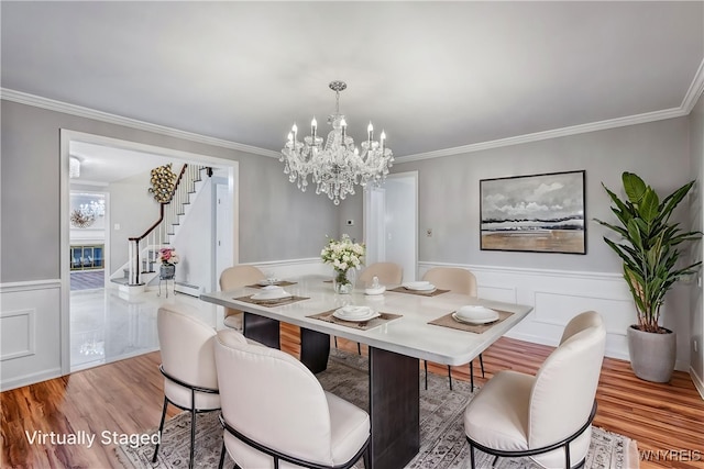 dining room featuring stairs, a chandelier, crown molding, a wainscoted wall, and light wood-style floors