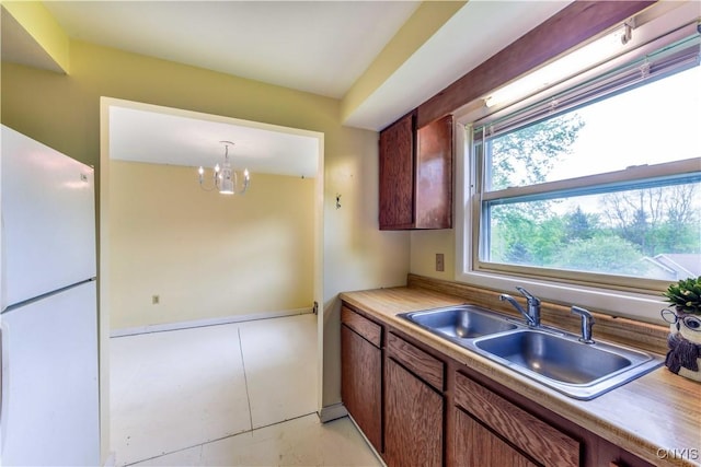 kitchen featuring freestanding refrigerator, a sink, a chandelier, light countertops, and brown cabinetry