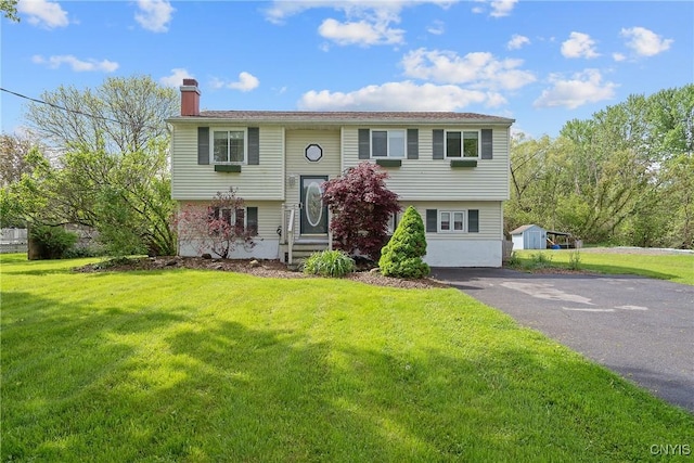 bi-level home featuring a chimney, a front lawn, entry steps, asphalt driveway, and a storage shed