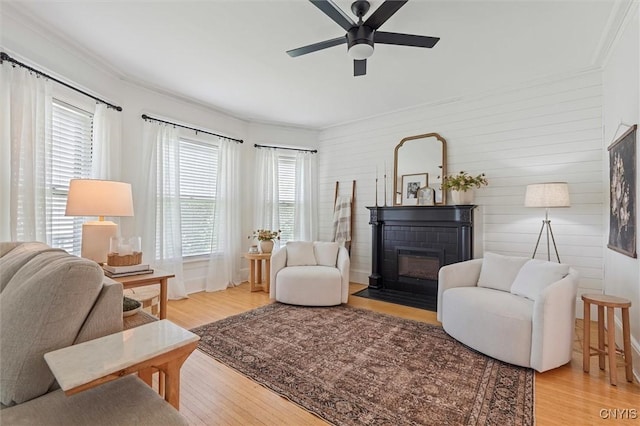 living area featuring light wood-style floors, a ceiling fan, a fireplace with flush hearth, ornamental molding, and wooden walls