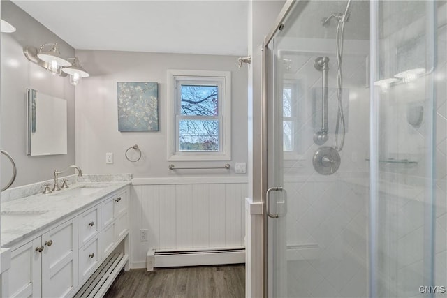 full bathroom with a baseboard radiator, a shower stall, wood finished floors, a wainscoted wall, and double vanity