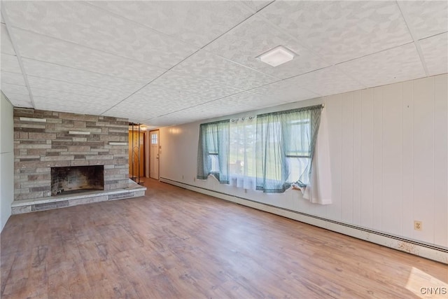 unfurnished living room featuring baseboard heating, wood finished floors, a stone fireplace, and a paneled ceiling