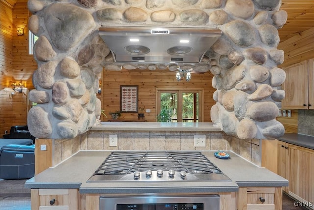 kitchen with light brown cabinets, oven, stainless steel gas stovetop, range hood, and wood walls