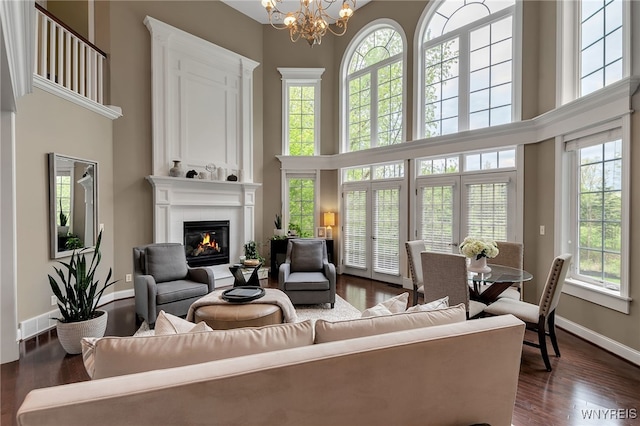 living room featuring a towering ceiling, a glass covered fireplace, a chandelier, dark wood-type flooring, and baseboards
