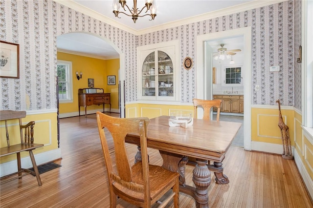 dining area with crown molding, wallpapered walls, light wood-style floors, arched walkways, and wainscoting