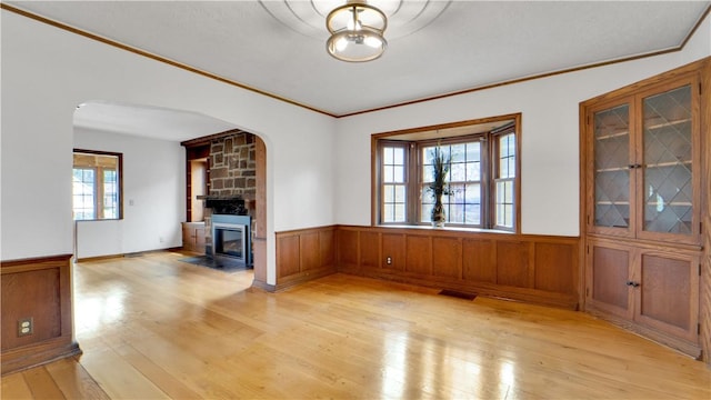unfurnished living room featuring light wood finished floors, crown molding, arched walkways, wainscoting, and a large fireplace
