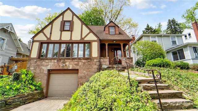tudor house with a garage, stone siding, driveway, stucco siding, and roof with shingles