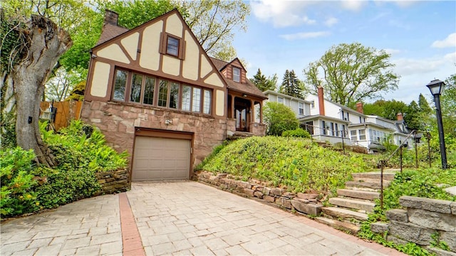 tudor home with a garage, stone siding, decorative driveway, a chimney, and stucco siding