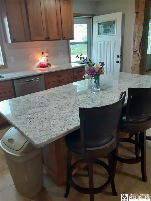 9165 Hebner Hill Rd, Otto, NY 14719 - kitchen with stainless steel dishwasher, brown cabinetry, a kitchen bar, and light stone counters