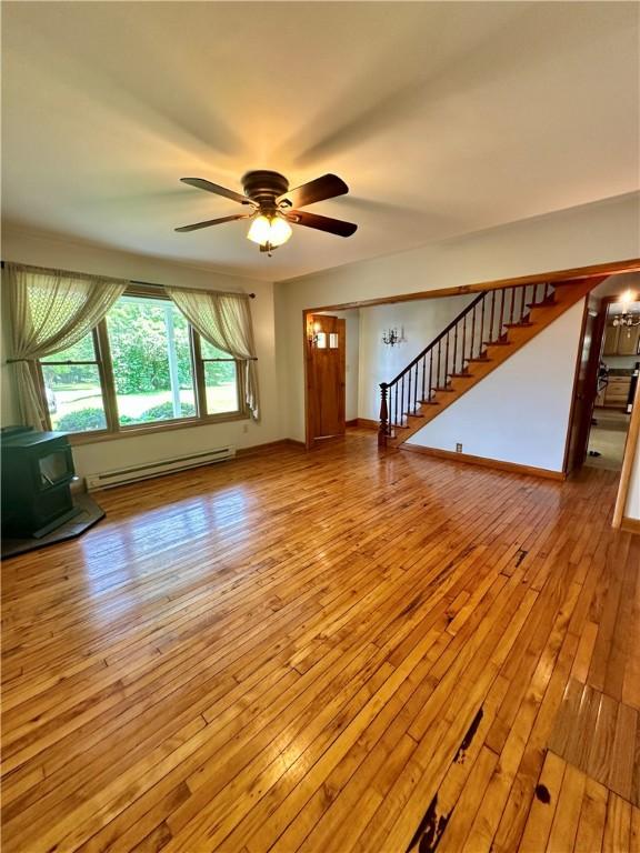 6956 E Washington St, Bath, NY 14810 - unfurnished living room featuring baseboard heating, a wood stove, wood-type flooring, stairs, and a ceiling fan