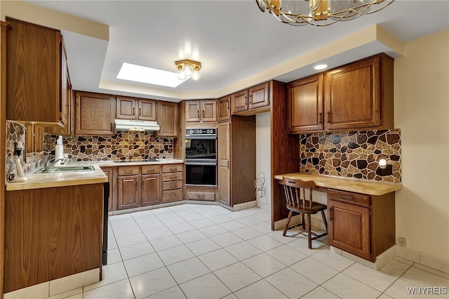 6182 Townline Rd, Wheatfield, NY 14094 - kitchen with under cabinet range hood, brown cabinetry, a chandelier, light countertops, and decorative backsplash