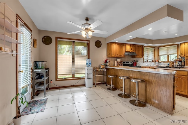 2160 Robert Dr, Niagara, NY 14304 - kitchen with stainless steel electric range, under cabinet range hood, a ceiling fan, brown cabinets, and healthy amount of natural light