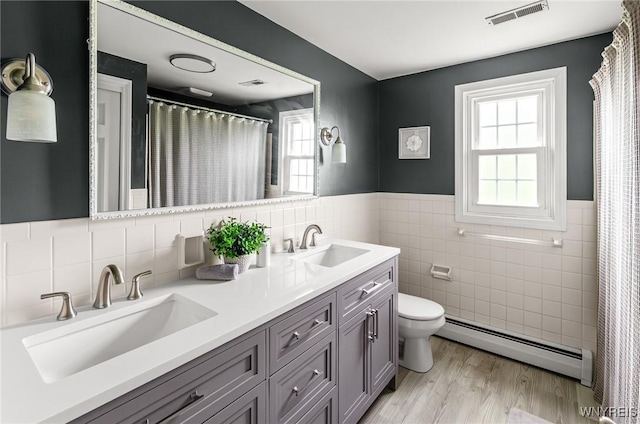 79 Hemlock Dr, West Seneca, NY 14224 - bathroom featuring baseboard heating, tile walls, wainscoting, double vanity, and wood finished floors
