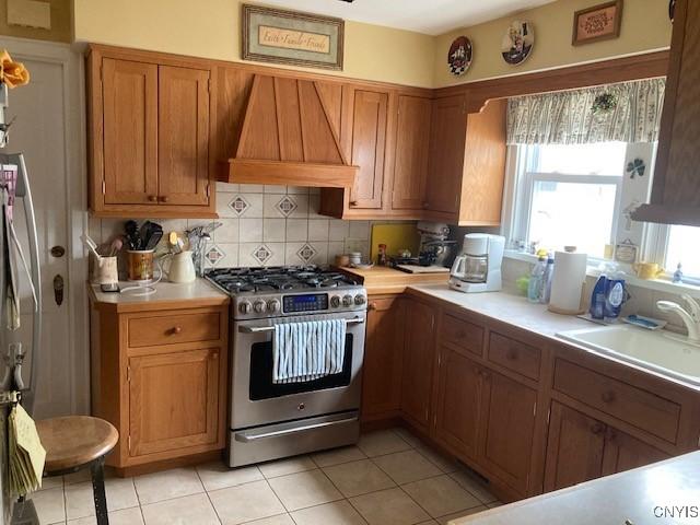 820 James St, Utica, NY 13501 - kitchen featuring stainless steel range with gas cooktop, custom exhaust hood, light countertops, brown cabinetry, and tasteful backsplash