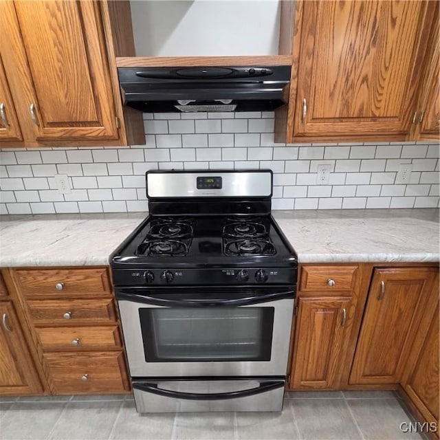 5709 Merry St, Verona, NY 13478 - kitchen featuring stainless steel gas stove, brown cabinetry, under cabinet range hood, light tile patterned flooring, and backsplash