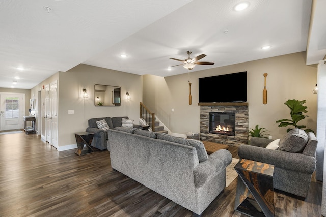 living area featuring a stone fireplace, a ceiling fan, dark wood-type flooring, baseboards, and stairs