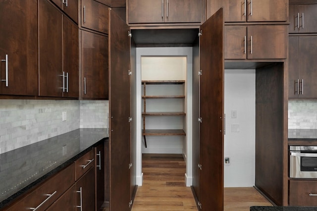 kitchen featuring tasteful backsplash, dark brown cabinets, oven, dark stone countertops, and light wood-type flooring