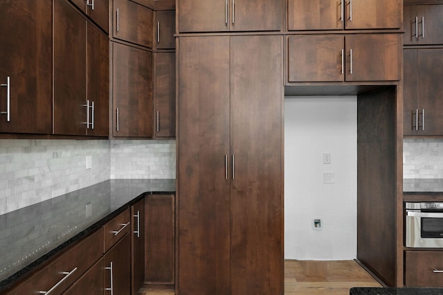 kitchen with dark brown cabinetry, oven, tasteful backsplash, dark stone counters, and light hardwood / wood-style flooring