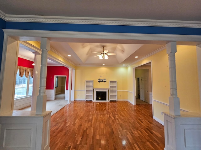 unfurnished living room featuring ceiling fan, wood-type flooring, a raised ceiling, ornamental molding, and decorative columns