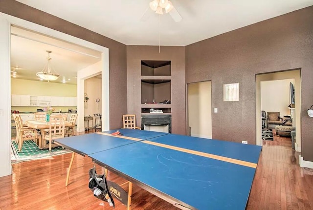 recreation room with ceiling fan, a fireplace, built in shelves, and wood-type flooring