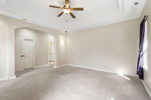 empty room featuring a tray ceiling, arched walkways, a ceiling fan, light carpet, and baseboards