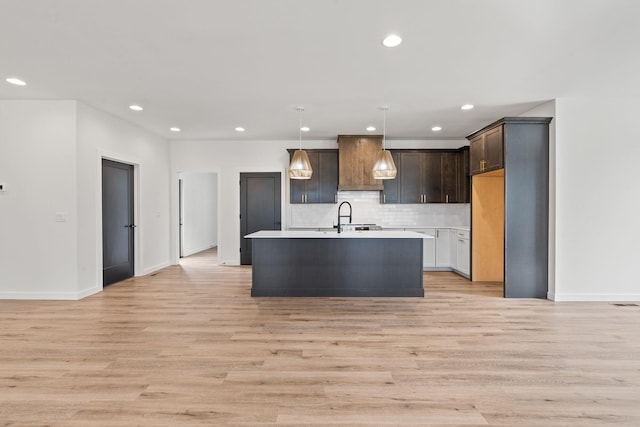 kitchen featuring an island with sink, hanging light fixtures, tasteful backsplash, dark brown cabinets, and light hardwood / wood-style floors