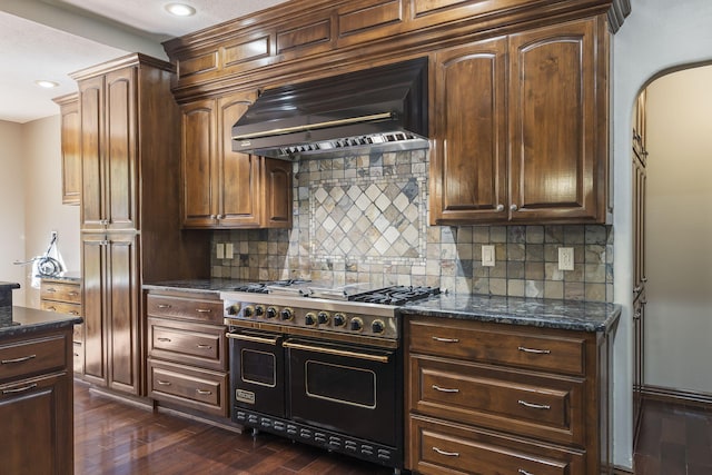 kitchen featuring decorative backsplash, dark wood-type flooring, dark stone counters, wall chimney range hood, and double oven range