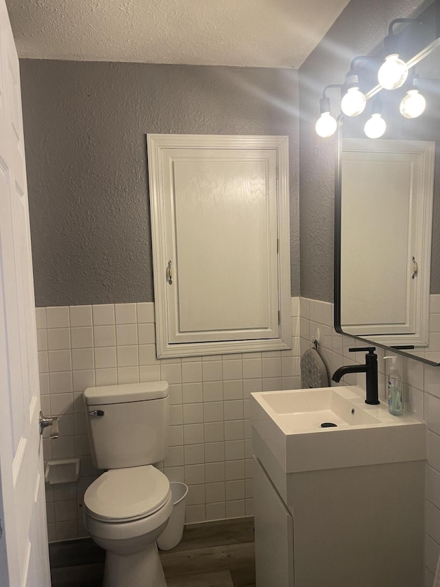 bathroom with a textured wall, toilet, vanity, tile walls, and wainscoting