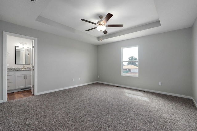 unfurnished bedroom featuring ceiling fan, a raised ceiling, ensuite bath, and dark colored carpet