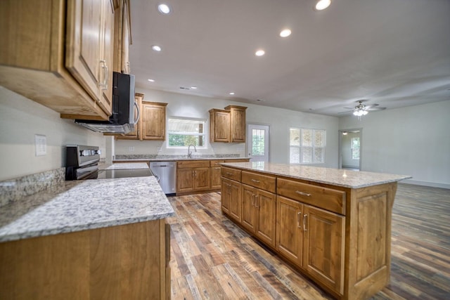 kitchen with ceiling fan, dark wood-type flooring, light stone counters, stainless steel appliances, and a center island