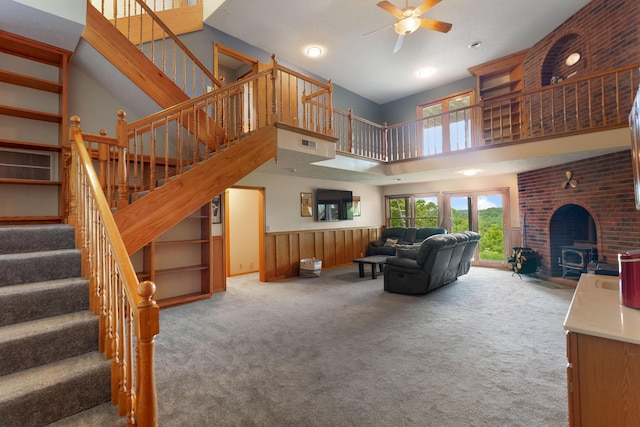 carpeted living room with visible vents, a towering ceiling, stairs, ceiling fan, and a wood stove