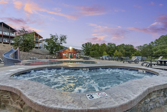 pool at dusk featuring a water slide, a patio, and a hot tub