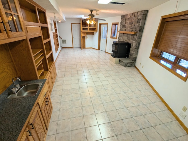 unfurnished living room with a sink, baseboards, ceiling fan, a wood stove, and light tile patterned floors