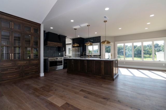 kitchen with an island with sink, custom range hood, dark wood-type flooring, premium appliances, and hanging light fixtures