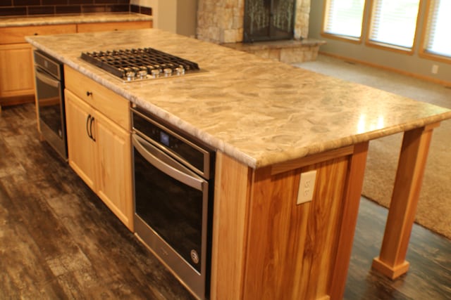 kitchen featuring a center island, a stone fireplace, light brown cabinets, dark wood-type flooring, and appliances with stainless steel finishes